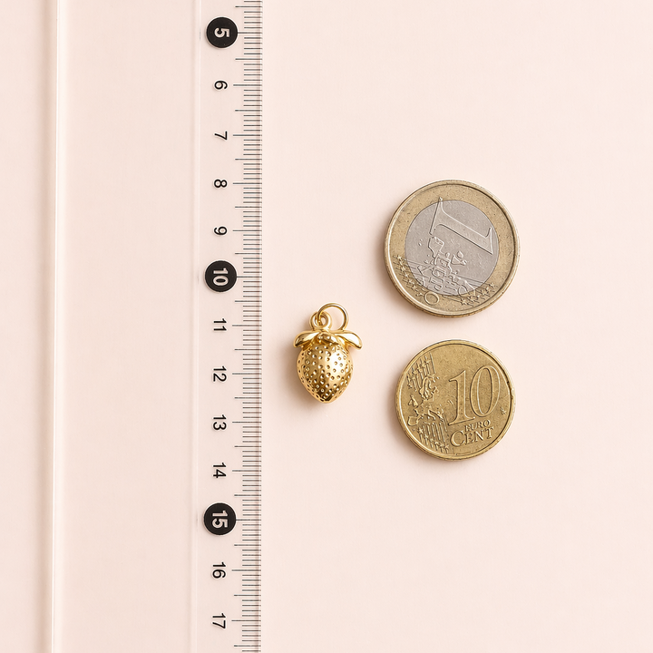 Gold strawberry charm next to two coins on a pink surface with a ruler for scale.