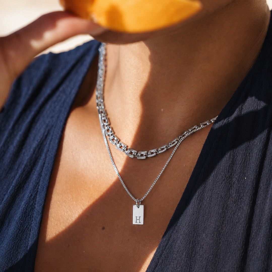 Person wearing layered silver necklaces with a visible 'H' pendant, holding an orange.