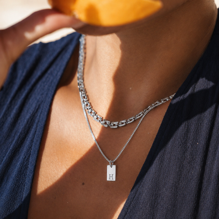 Person wearing layered silver necklaces with a visible 'H' pendant, holding an orange.