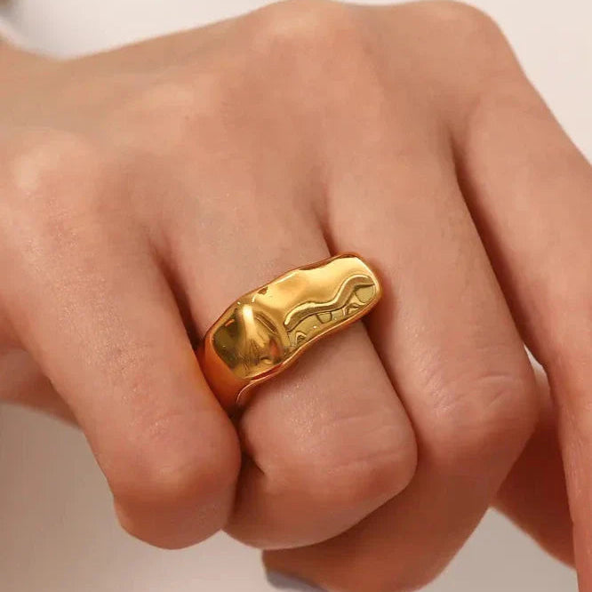 Gold chunky ring on a person's finger against a neutral background
