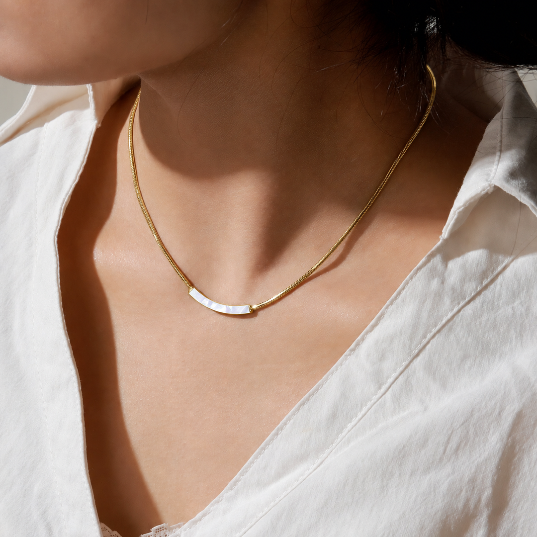 A close-up of a model wearing a delicate gold chain necklace with a small curved white inlay at the center, styled with a white collared shirt.