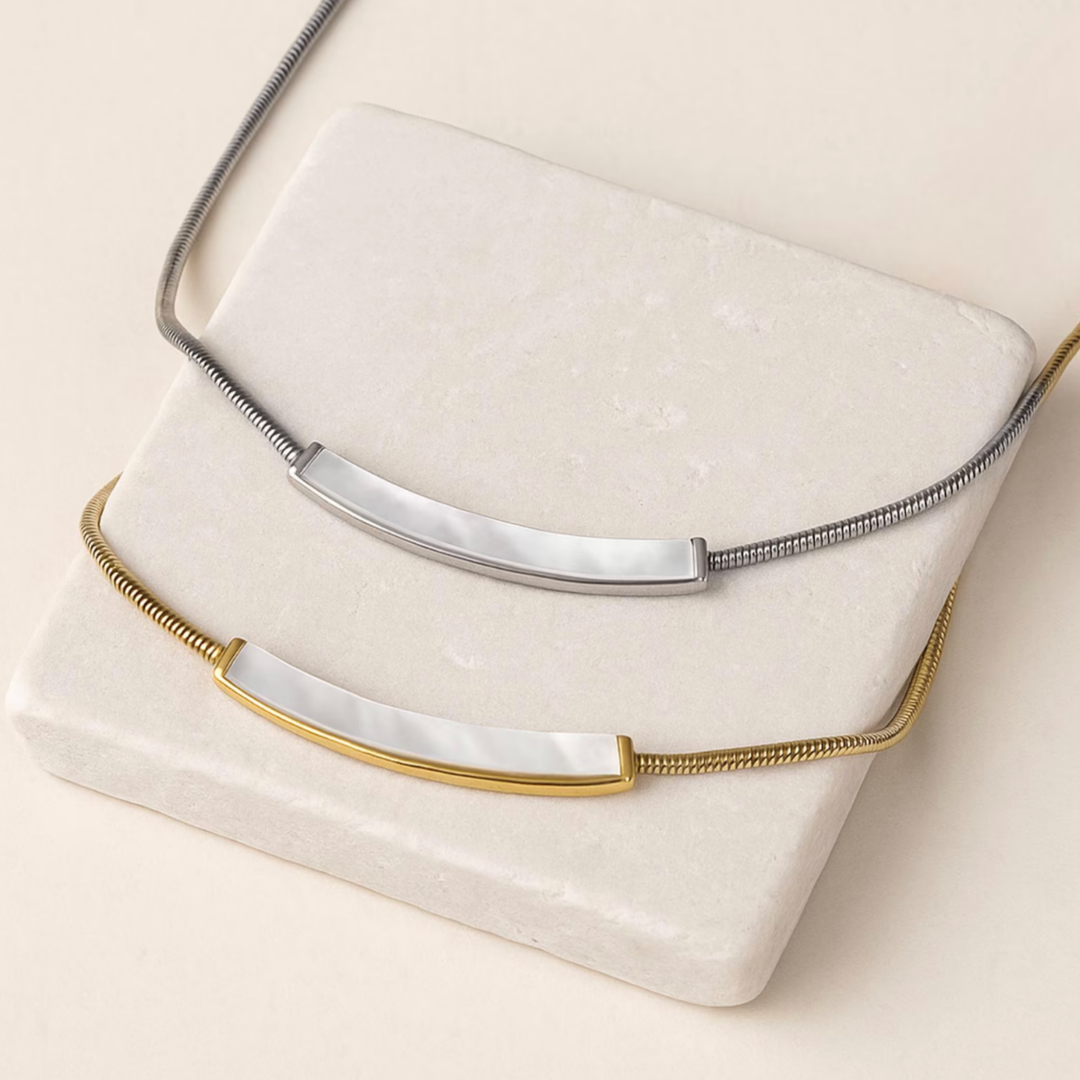 Two minimalist bar nameplates displayed on a cream stone block, one with a silver snake chain and one with a gold snake chain.