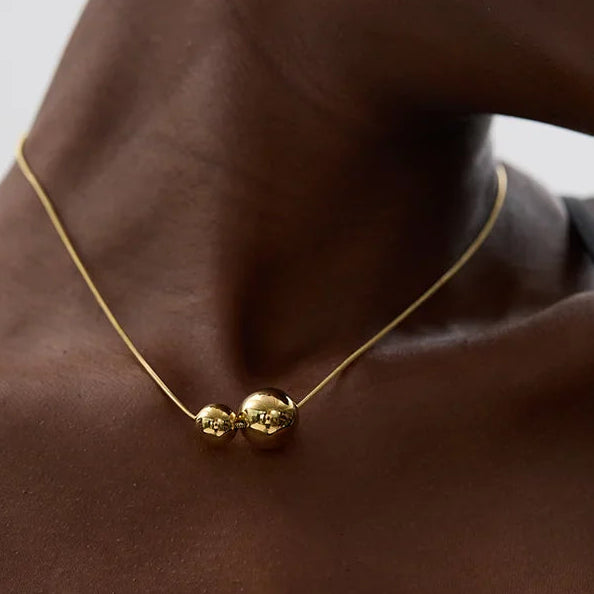 Gold necklace with two spherical pendants on a person's neck against a neutral background