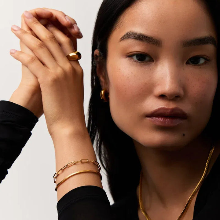Woman wearing gold jewelry including a ring, bracelet, and necklace on a neutral background