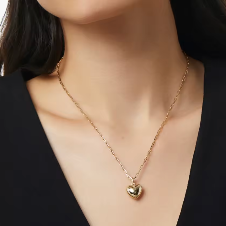 A close-up of a model wearing a delicate gold chain necklace with a small polished gold heart pendant, styled with a black top.