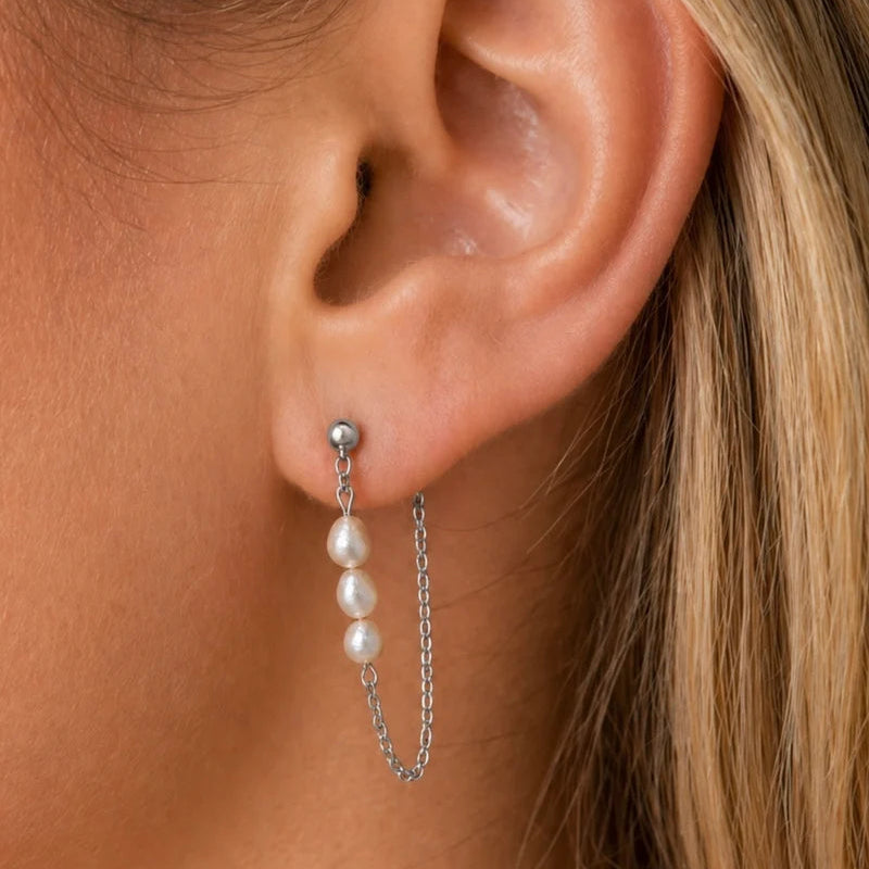 A close-up of a model's ear wearing a silver stud earring with three dangling white pearls and a short chain.