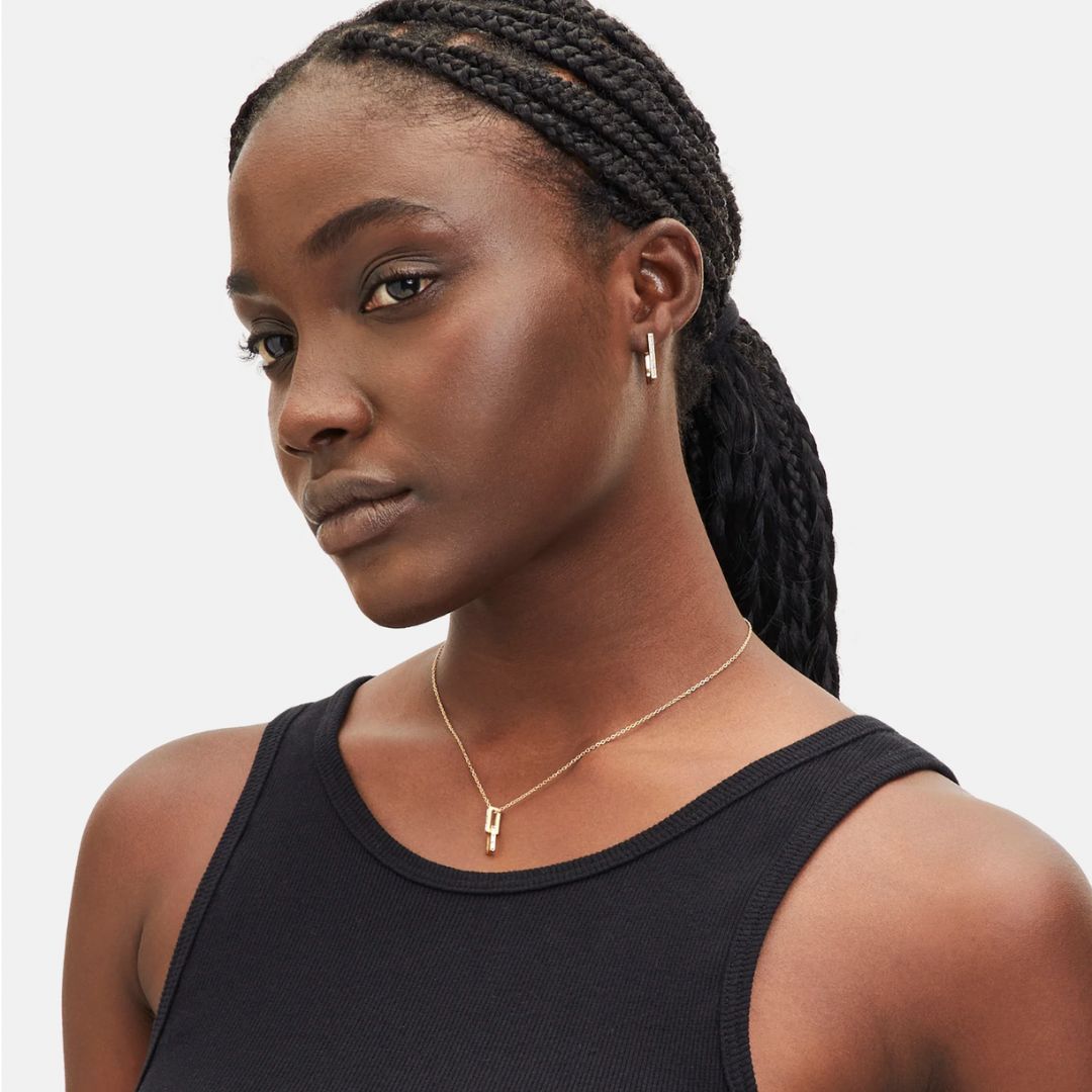 Woman with braided hair wearing a black tank top and gold necklace on a light gray background