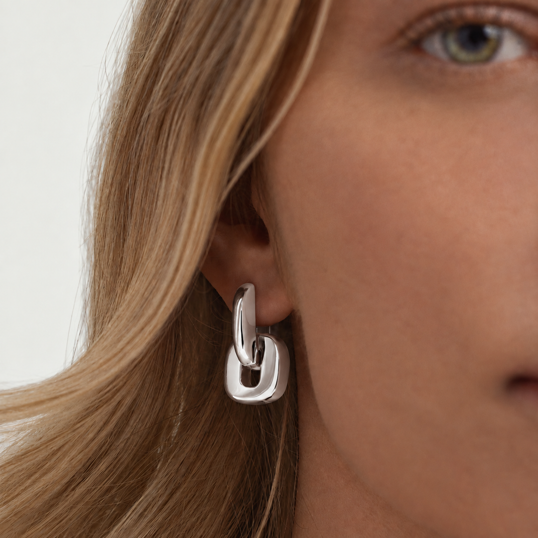 A close-up of a model's ear wearing a polished silver geometric hoop earring with a thick interlocking link design.