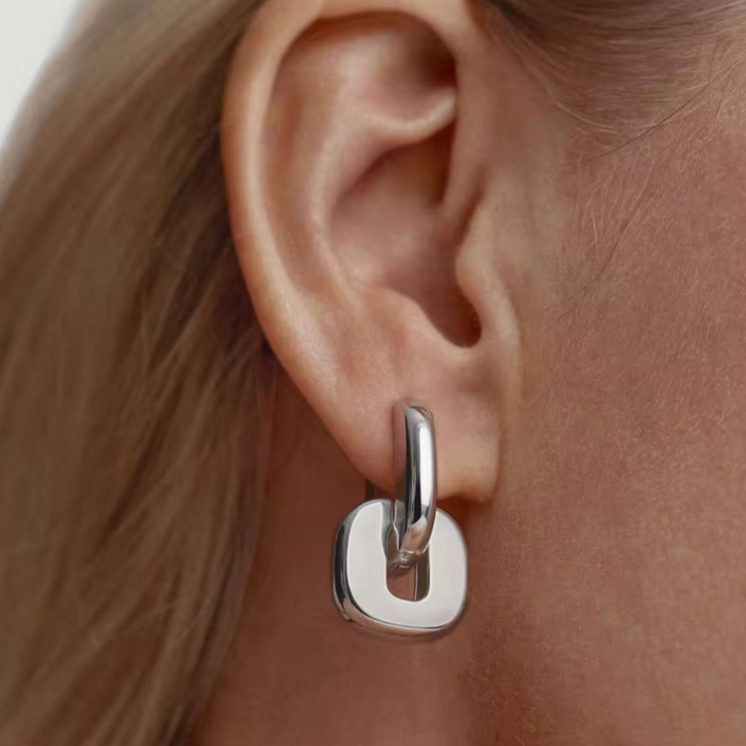A close-up of a person's ear wearing a polished silver huggie hoop earring with a square drop pendant.