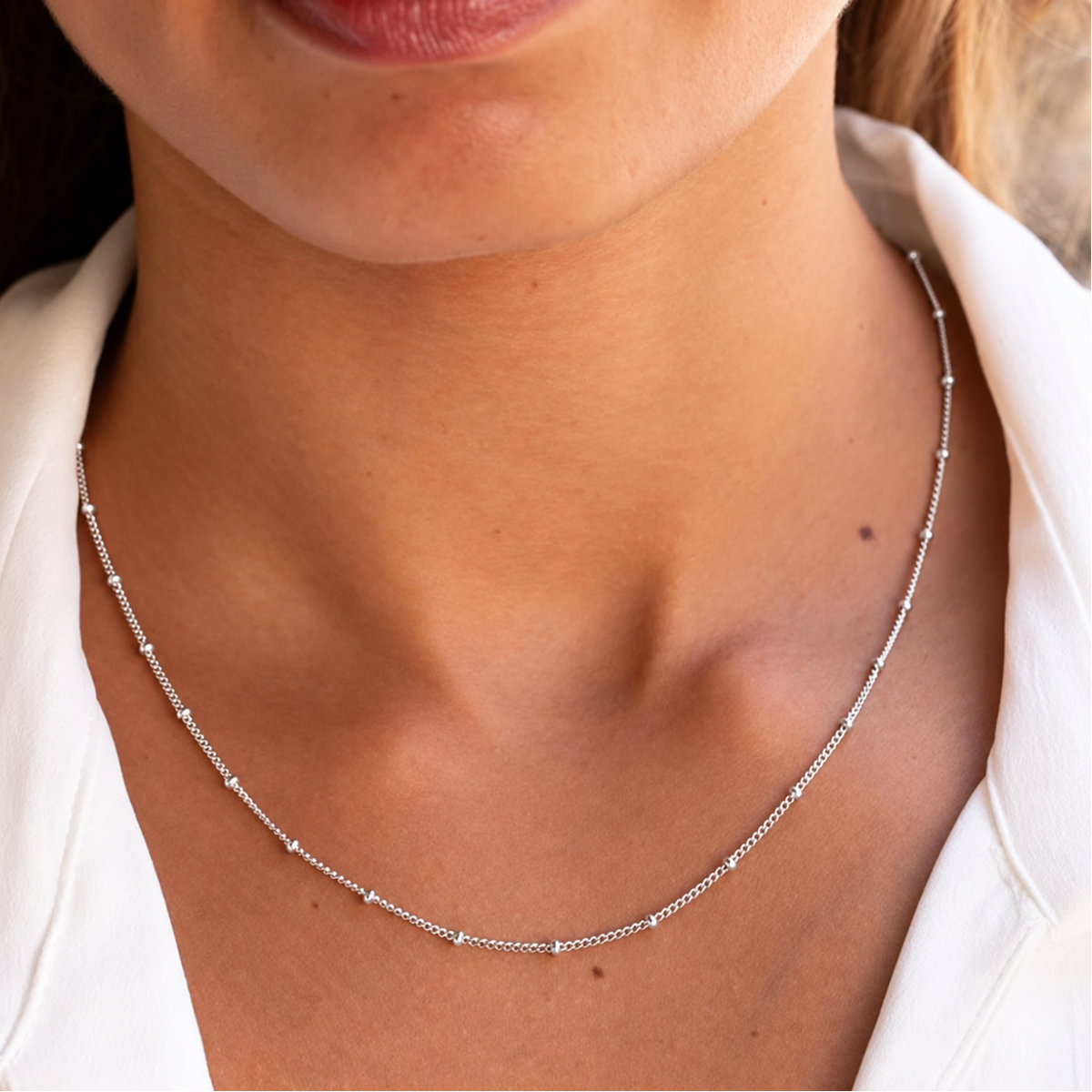 A close-up of a woman's neck and collarbone wearing a delicate silver beaded chain necklace with small clear stones, paired with a white collared shirt.