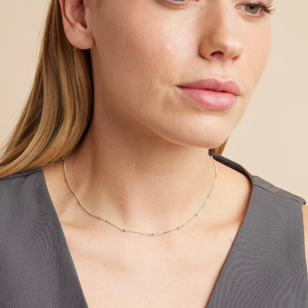 A close-up of a woman wearing a delicate silver chain necklace with small star-shaped beads, paired with a dark gray sleeveless top against a warm beige background.
