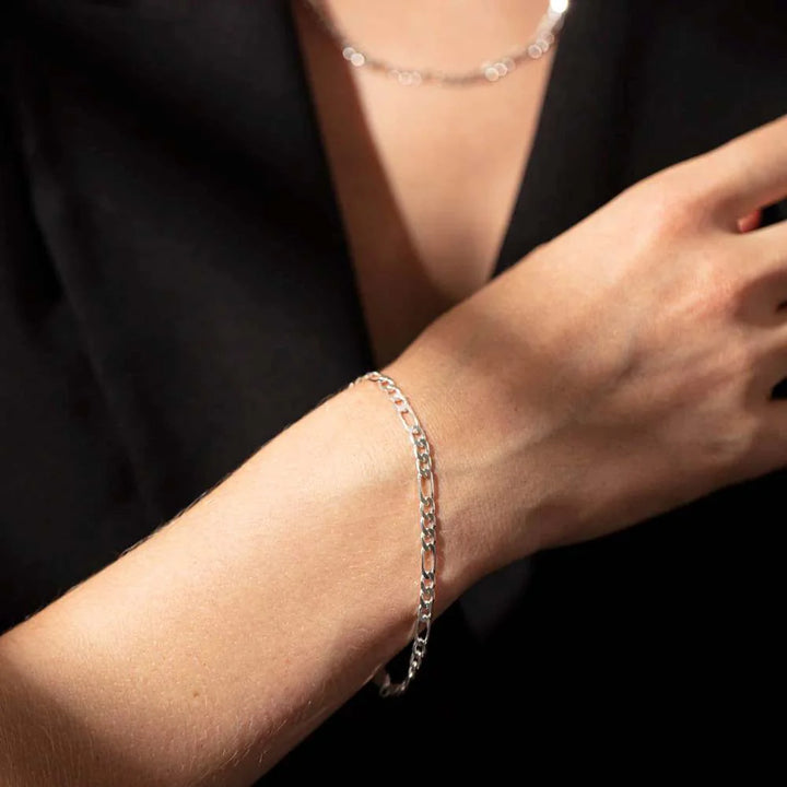 A close-up of a person's forearm wearing a delicate silver curb chain bracelet, paired with a black top and a matching silver necklace.