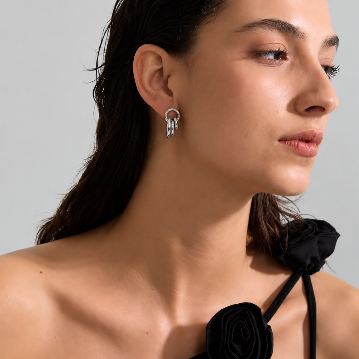 A close-up of a woman wearing a silver hoop earring with three dangling curved silver bars. She is wearing a black halter top with fabric roses, set against a plain light gray background.