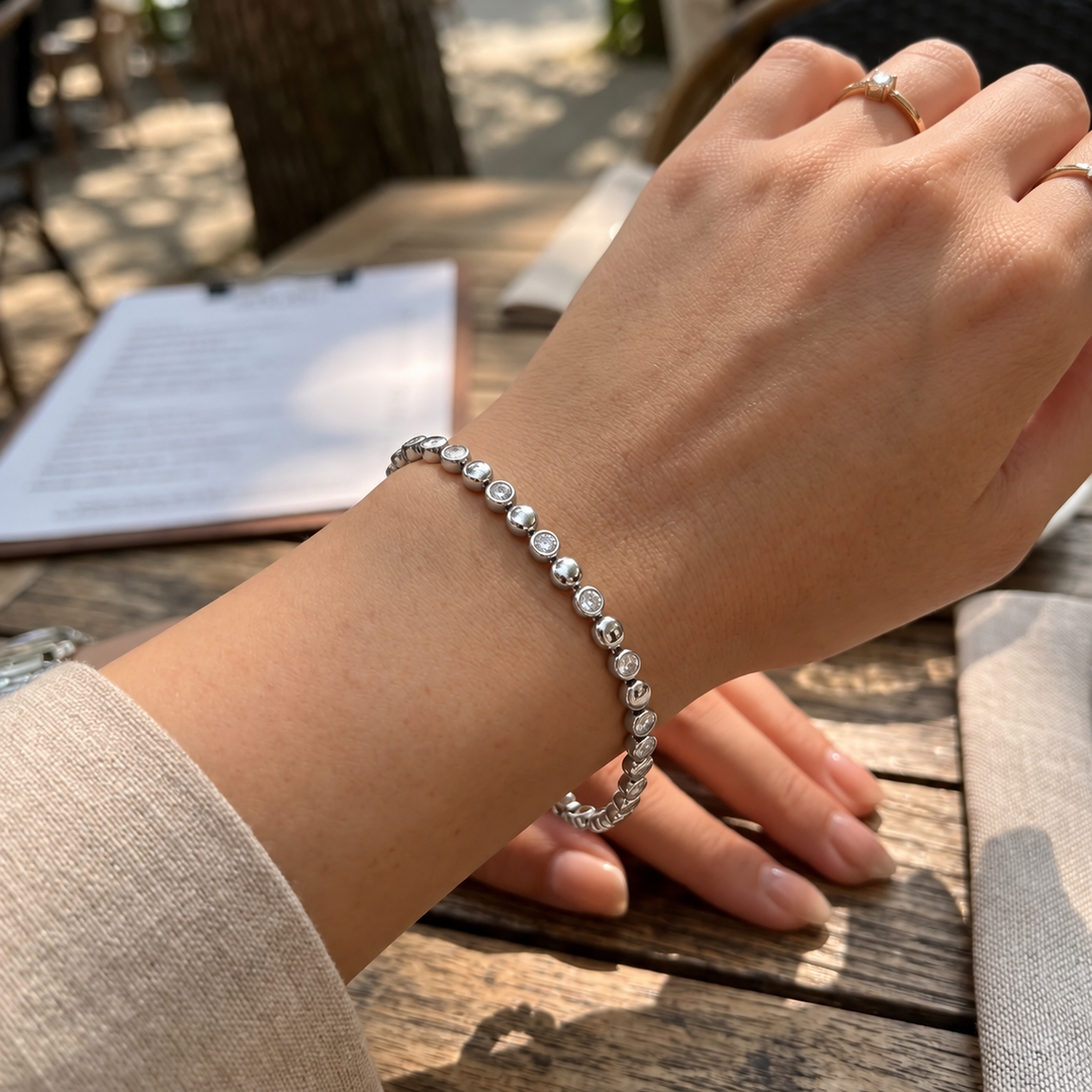 A close-up lifestyle shot of a person's wrist wearing a delicate silver beaded bracelet with clear round stones, set against a blurred outdoor cafe background.