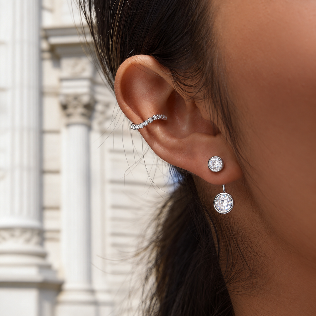A close-up of a person's ear wearing a silver ear cuff with small clear stones and a matching silver stud earring with a round clear stone, set against a blurred white classical building background.