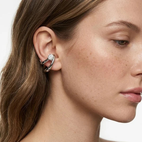 A close-up of a woman's ear wearing a silver-toned curved hoop earring with a small clear gemstone, shown against a light neutral background.