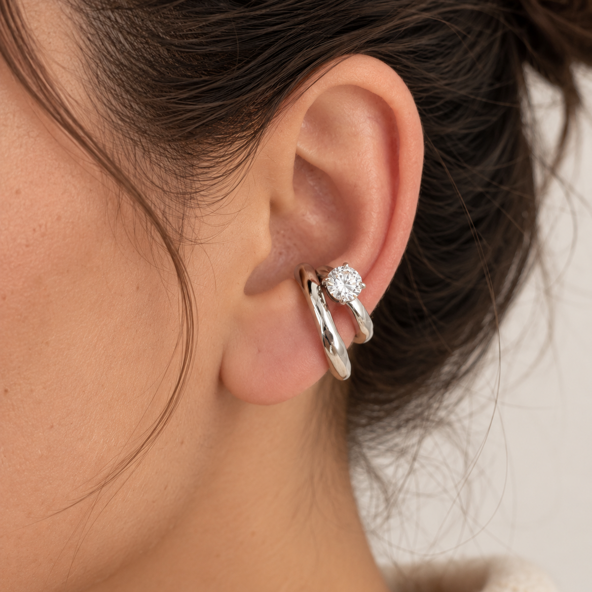 A close-up of a person's ear wearing a silver huggie hoop earring with a small round clear gemstone, shown against a soft light background.