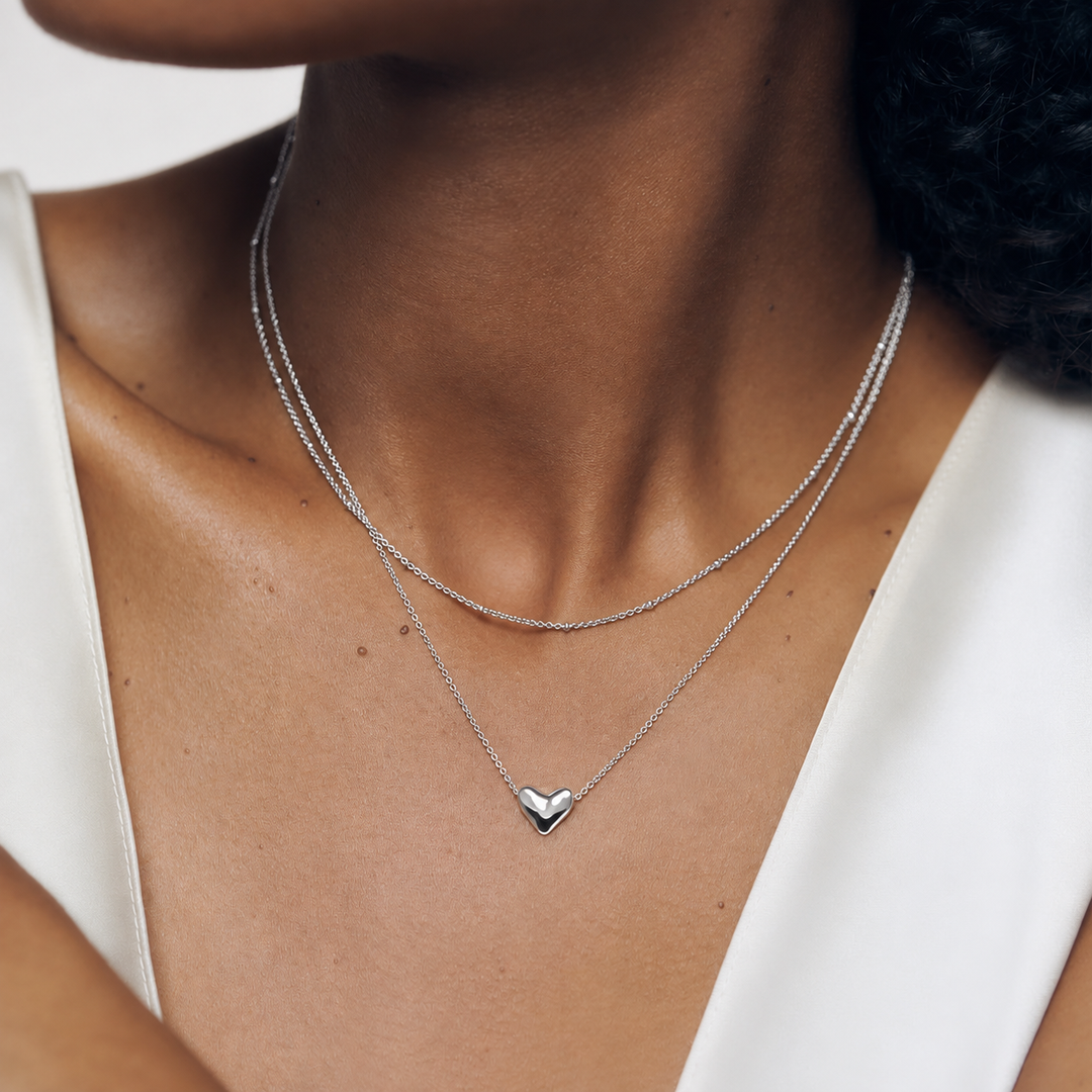 A close-up of a model wearing a delicate double-layered silver necklace with a small heart pendant at the center.