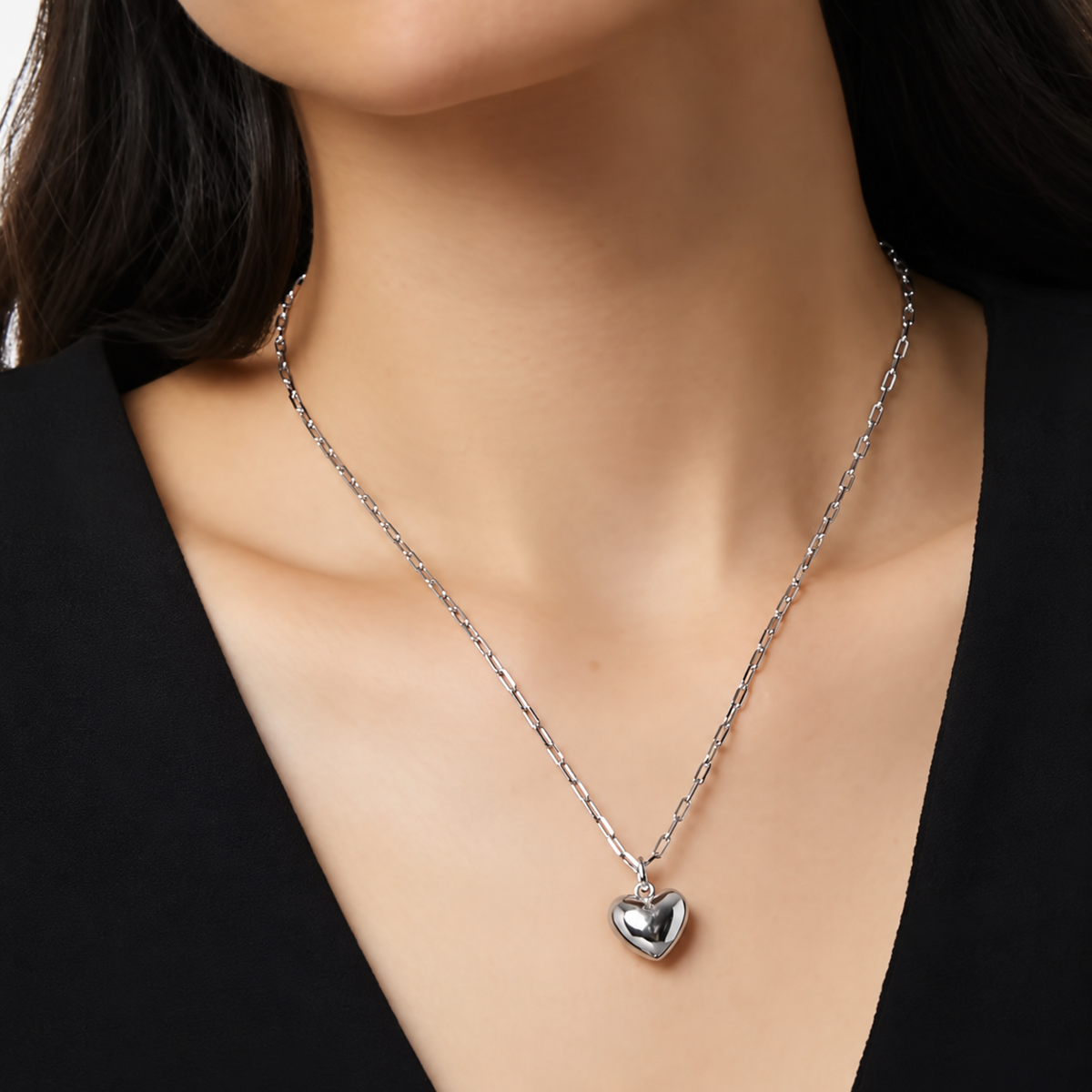A close-up of a model wearing a delicate silver chain necklace with a polished heart-shaped pendant, styled with a black top.