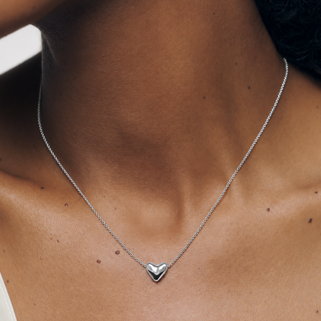 A close-up of a person's neck and upper chest wearing a delicate silver chain necklace with a small polished heart pendant.