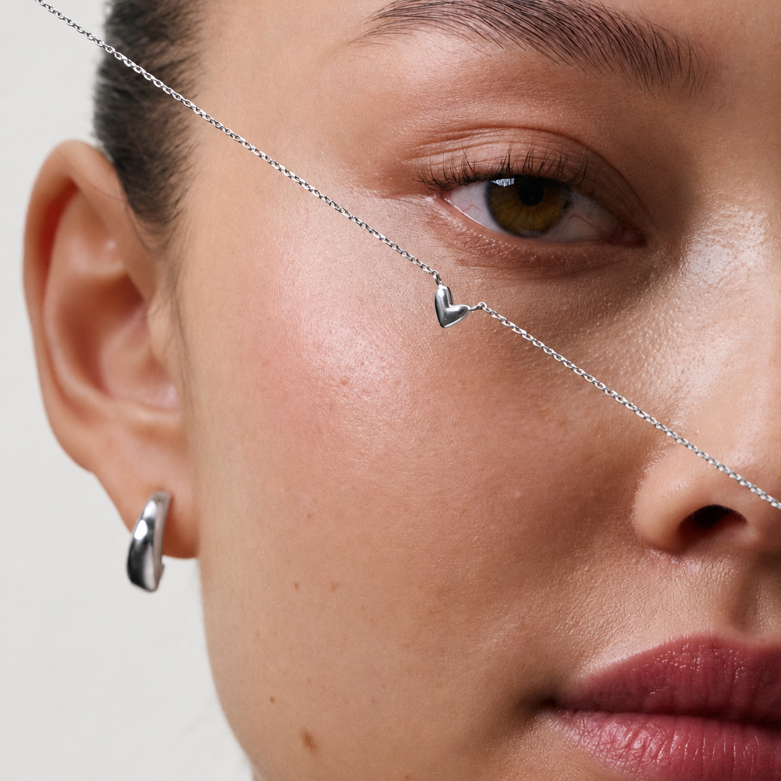 Close-up of a model wearing a delicate silver chain necklace with a small V-shaped pendant and a silver hoop earring against a light neutral background.