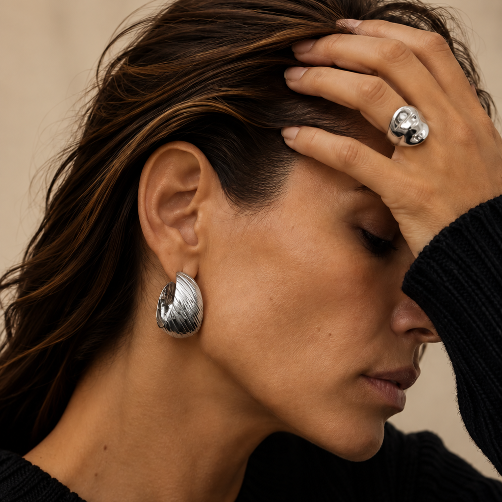 A close-up lifestyle shot of a woman with dark hair wearing a textured silver hoop earring and a matching silver ring on her finger, dressed in a black knit sweater against a neutral beige background.