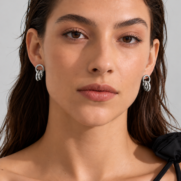 A close-up portrait of a woman wearing silver hoop earrings with a textured, layered circular design. The background is a clean, neutral light gray studio backdrop.