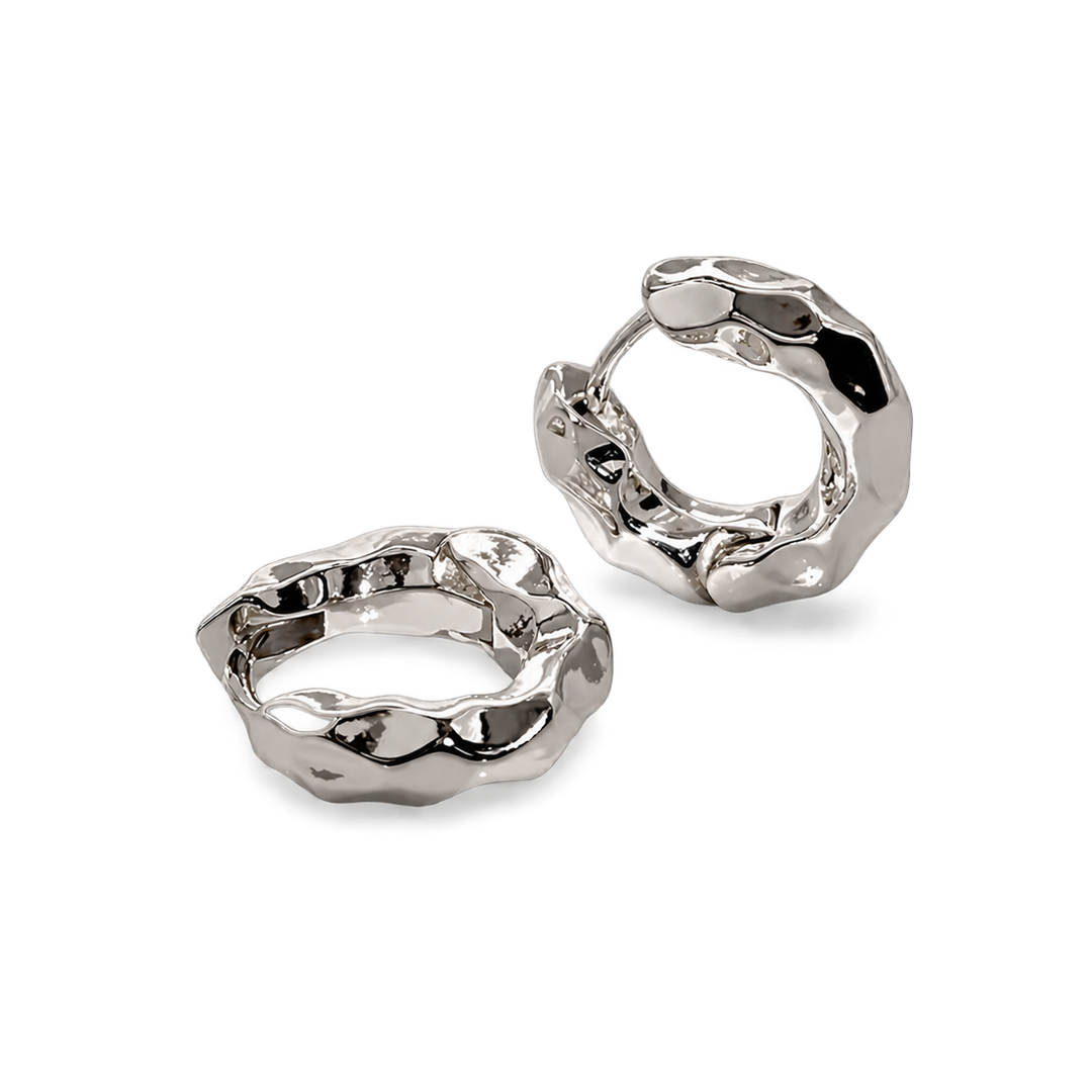 A pair of chunky, textured silver huggie hoop earrings with a polished, organic, hammered finish, displayed on a clean white background.