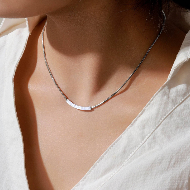 A close-up of a model's neck and chest wearing a delicate silver necklace with a curved, textured bar pendant, styled with a white blouse.