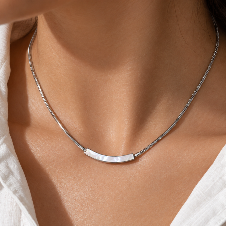 A close-up of a woman's neck and chest wearing a silver snake chain necklace with a curved bar pendant made of iridescent mother-of-pearl.