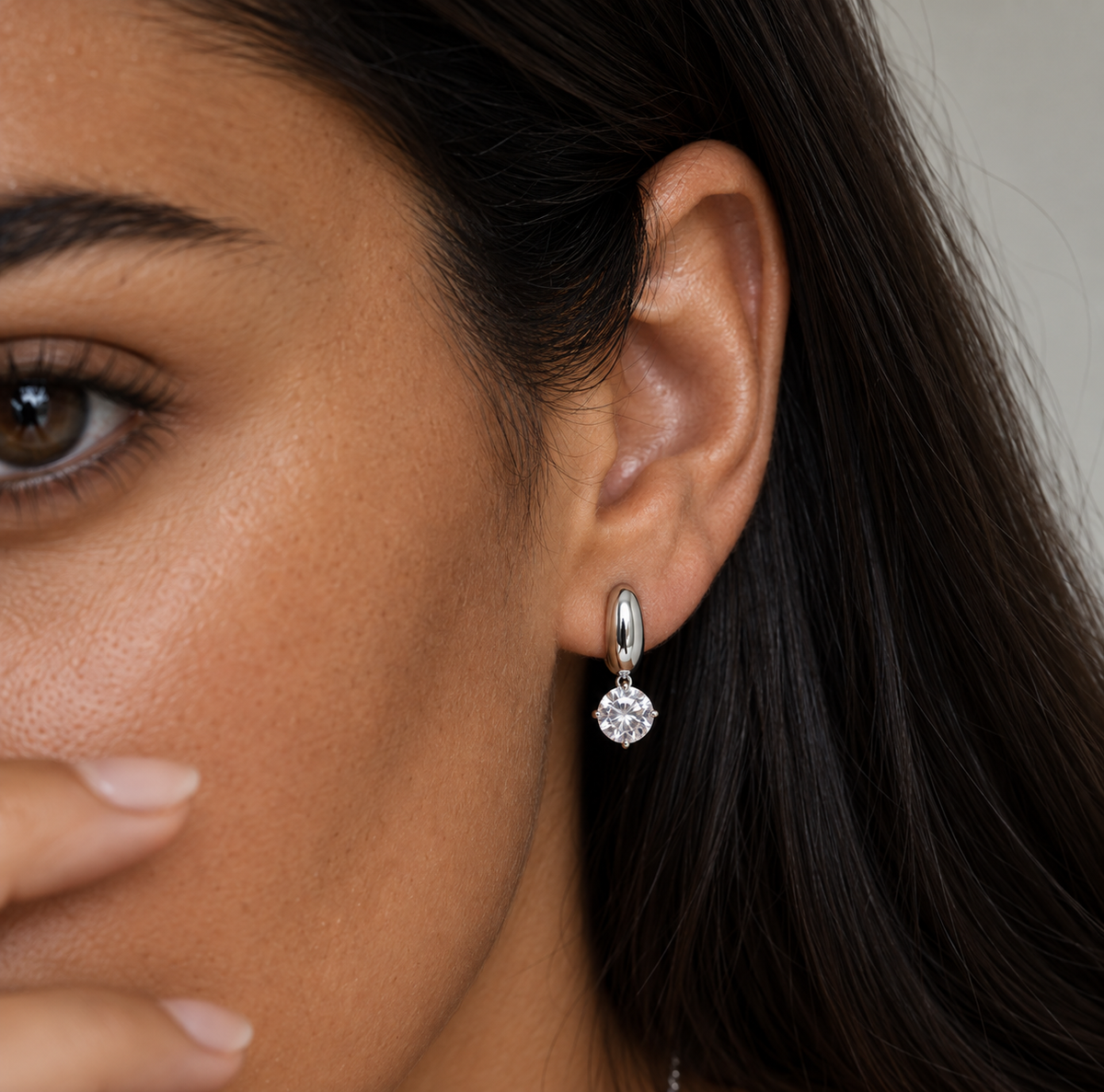 A close-up of a woman's ear wearing a small silver hoop earring with a clear round-cut gemstone.