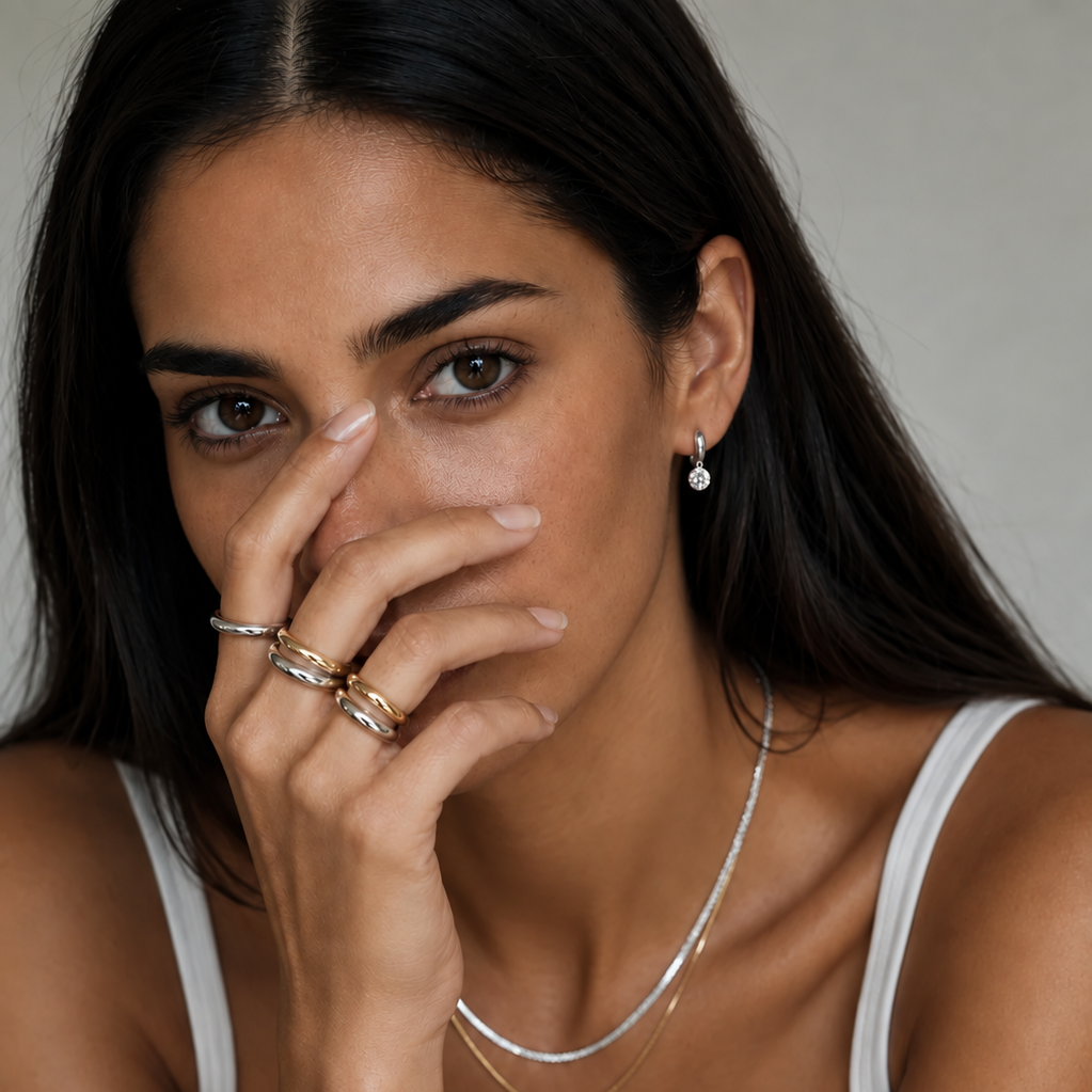 A close-up lifestyle shot of a woman wearing delicate layered jewelry, including a thin silver chain necklace, a gold bangle bracelet, and stacked rings on her fingers, paired with a black and white tank top.