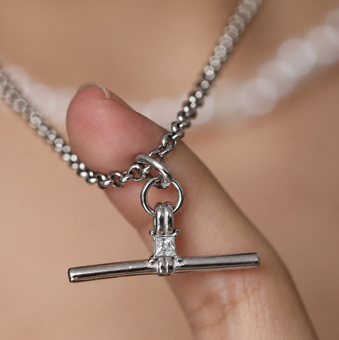 A close-up of a silver chain necklace with a horizontal bar pendant held by a person's fingers against a soft-focus skin background.
