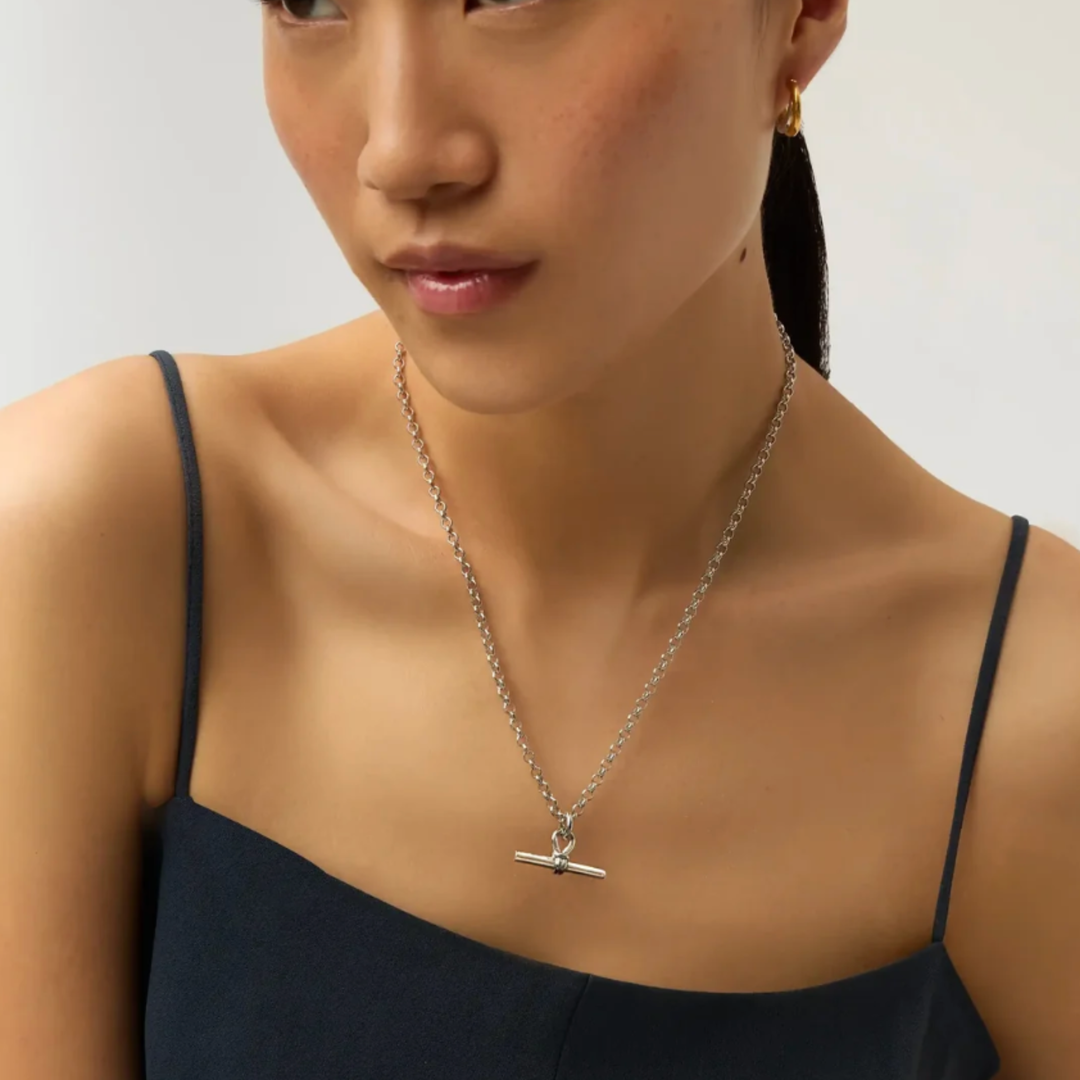 A close-up of a model wearing a delicate silver chain necklace with a horizontal bar toggle clasp pendant, paired with a dark navy camisole top.