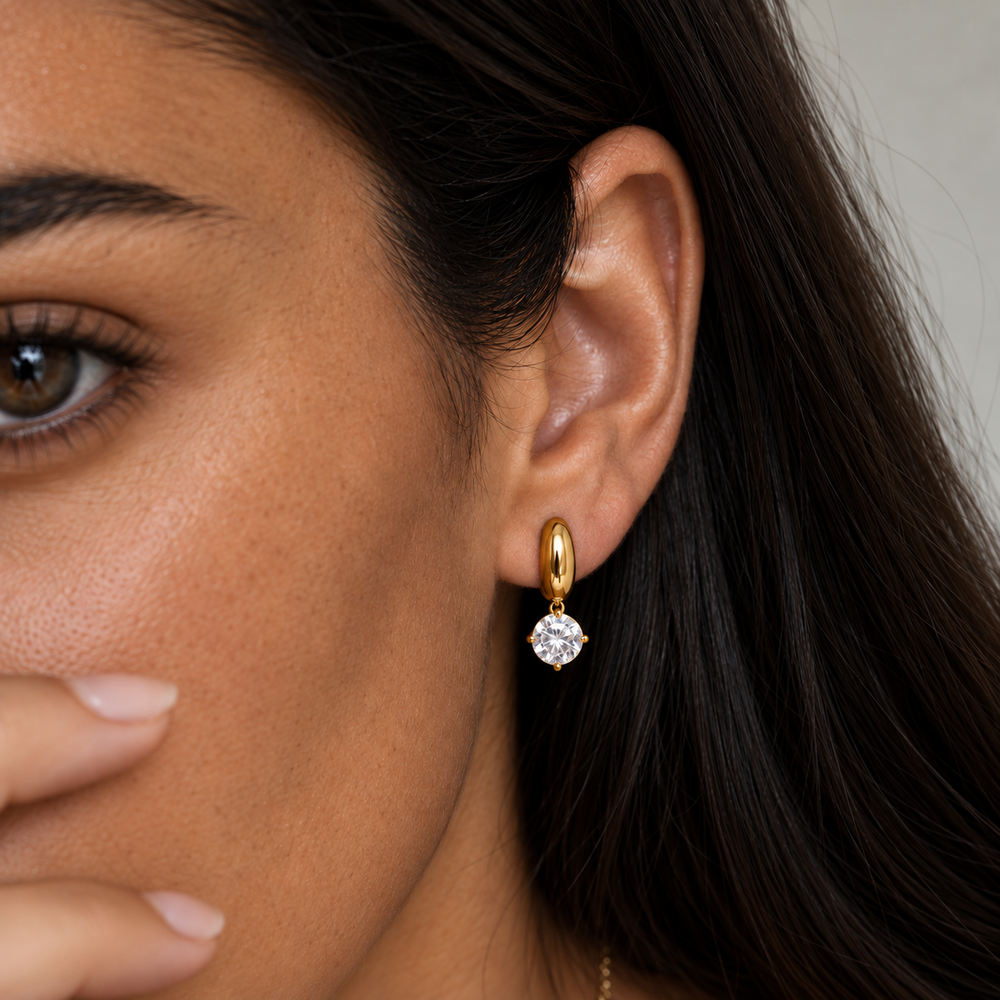A close-up of a woman's ear wearing a small gold huggie hoop earring with a clear round-cut gemstone dangling at the lobe.