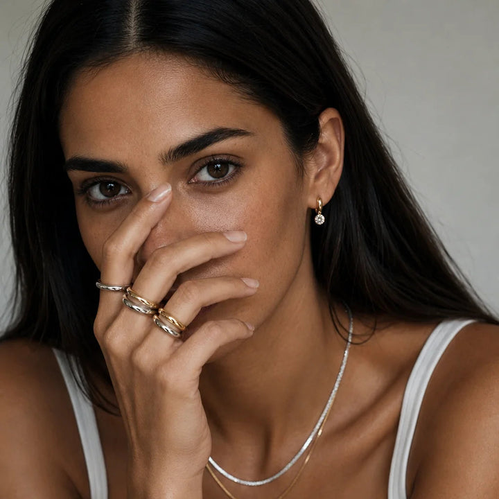 A close-up lifestyle shot of a woman wearing delicate layered jewelry, including a thin silver chain necklace, a gold bangle bracelet, and stacked rings on her fingers, while wearing a black and white tank top.