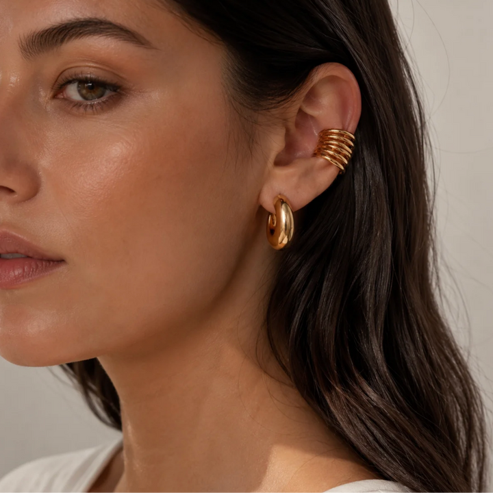 A close-up of a woman's ear wearing a gold ear cuff with stacked spiral bands and a small gold hoop earring, against a soft light gray background.