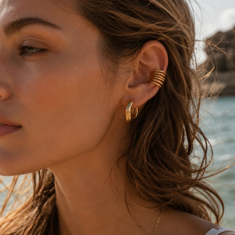 A close-up of a woman's ear wearing a stack of gold ear cuffs, including a textured spiral cuff and a small gold hoop, with a blurred outdoor background of water and rocks.