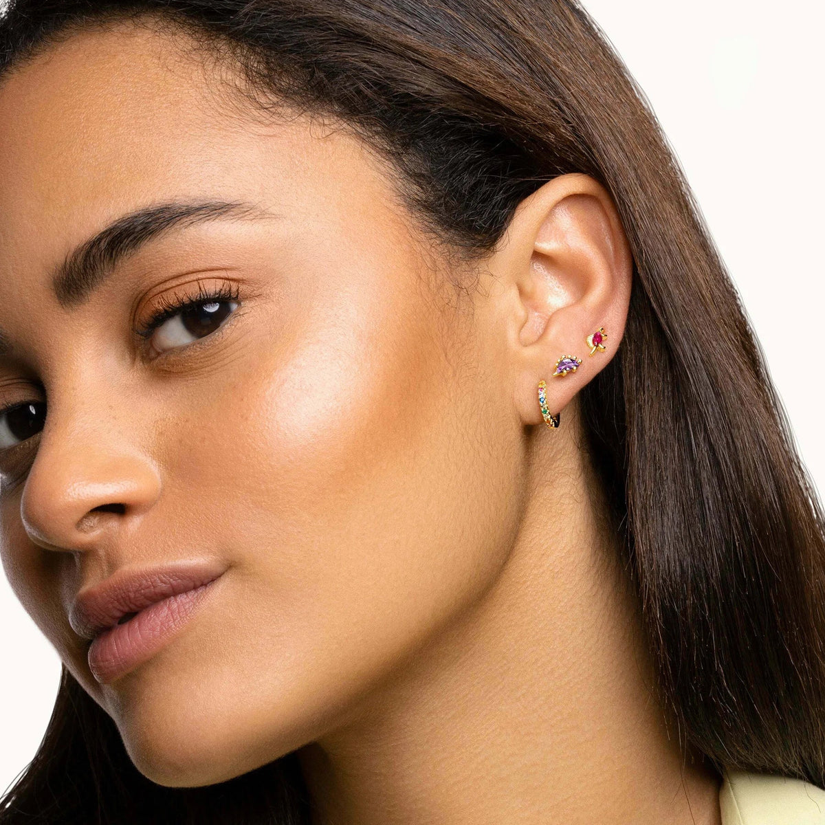 A close-up of a woman's ear wearing multiple small gold hoop and stud earrings, including one with colorful gemstones, against a clean white background.