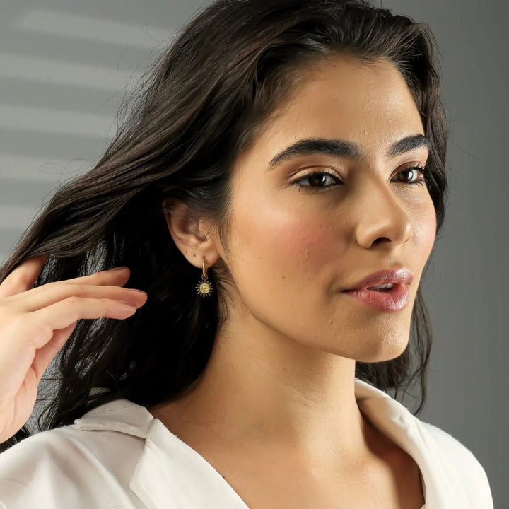 Woman with dark hair and earrings, wearing a white top against a neutral background