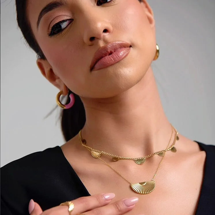 Woman wearing gold jewelry including a necklace, earrings, and ring on a neutral background