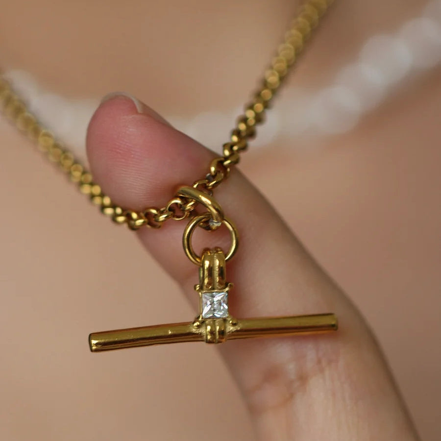 A close-up of a gold necklace with a toggle bar pendant held by a person's fingers against a soft beige skin background.
