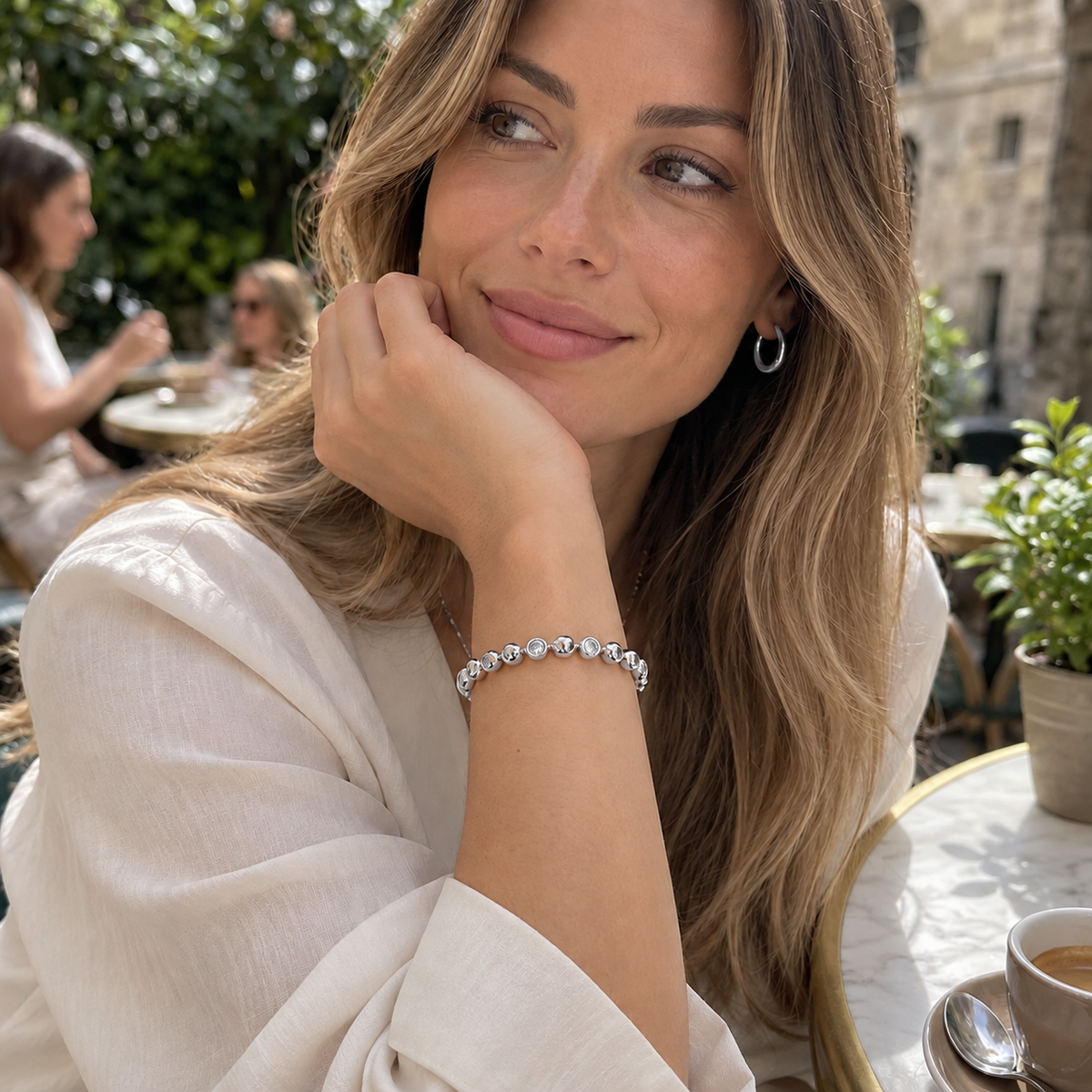 A woman with long light brown hair rests her chin on her hand at an outdoor cafe, wearing a delicate silver bracelet with clear round gemstones and a small silver hoop earring.
