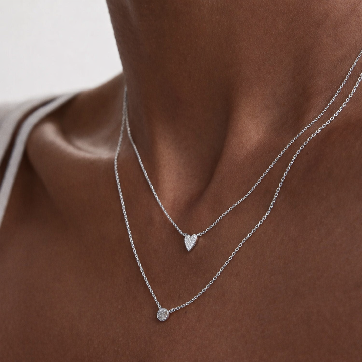 Silver necklaces with heart and round pendants on a person's neck against a neutral background