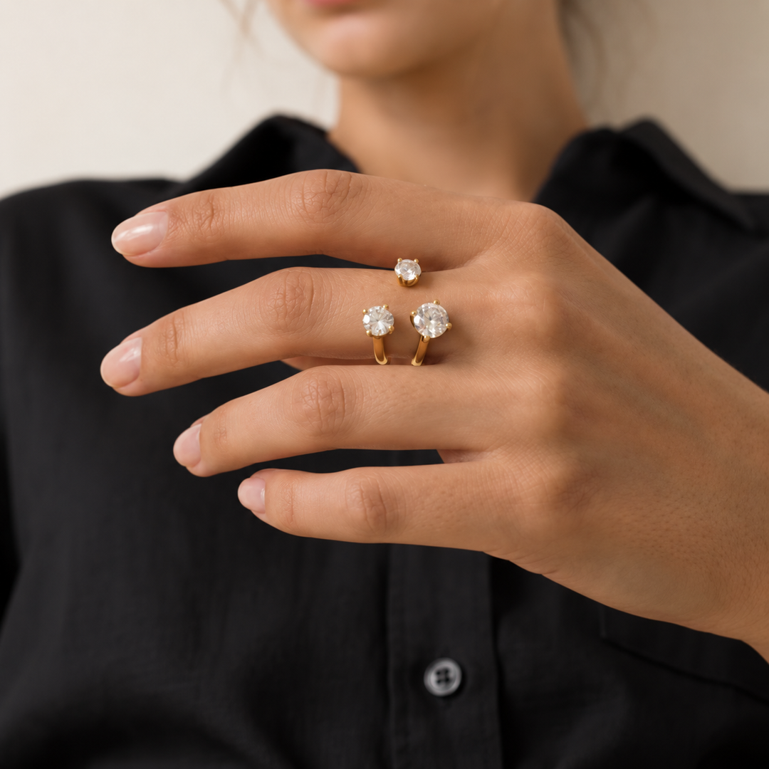 A close-up of a person's hand wearing three gold rings with clear gemstones, set against a black collared shirt and a soft beige background.