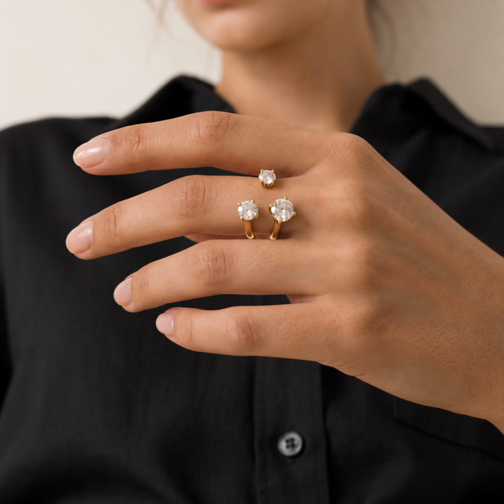 A close-up of a person's hand wearing three gold rings with clear gemstones, set against a black collared shirt and a soft beige background.