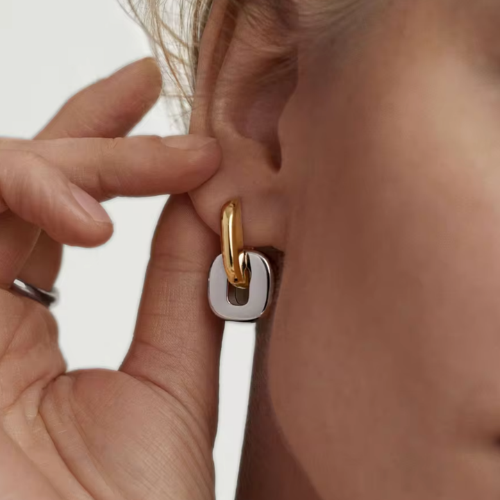 A close-up of a person's ear wearing a two-tone geometric hoop earring with a gold top loop and a silver square-shaped drop, while a hand adjusts it.
