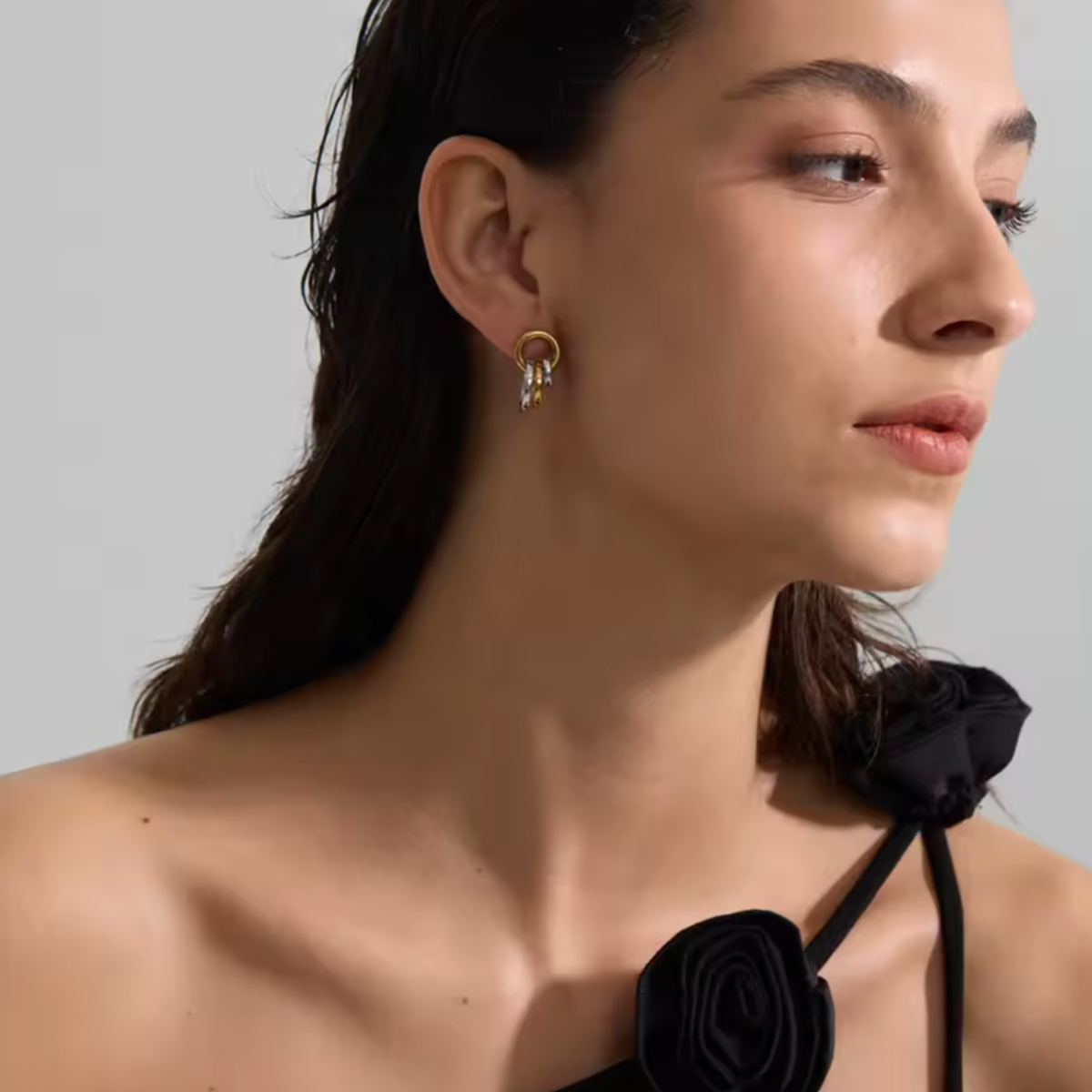 A close-up side profile of a woman wearing a delicate gold hoop earring with three small dangling clear stones, paired with a black rose-neck top against a light gray background.