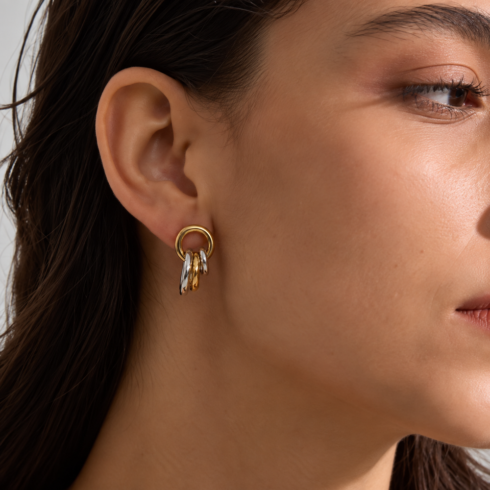 A close-up of a model's ear wearing a gold hoop earring with three dangling textured gold and silver leaf-like charms.