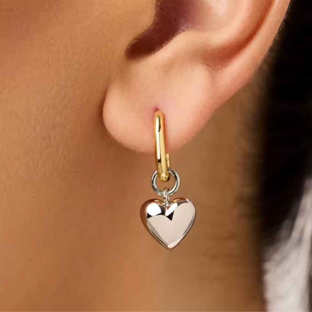 A close-up of a person's ear wearing a small gold hoop earring with a dangling polished silver heart charm.